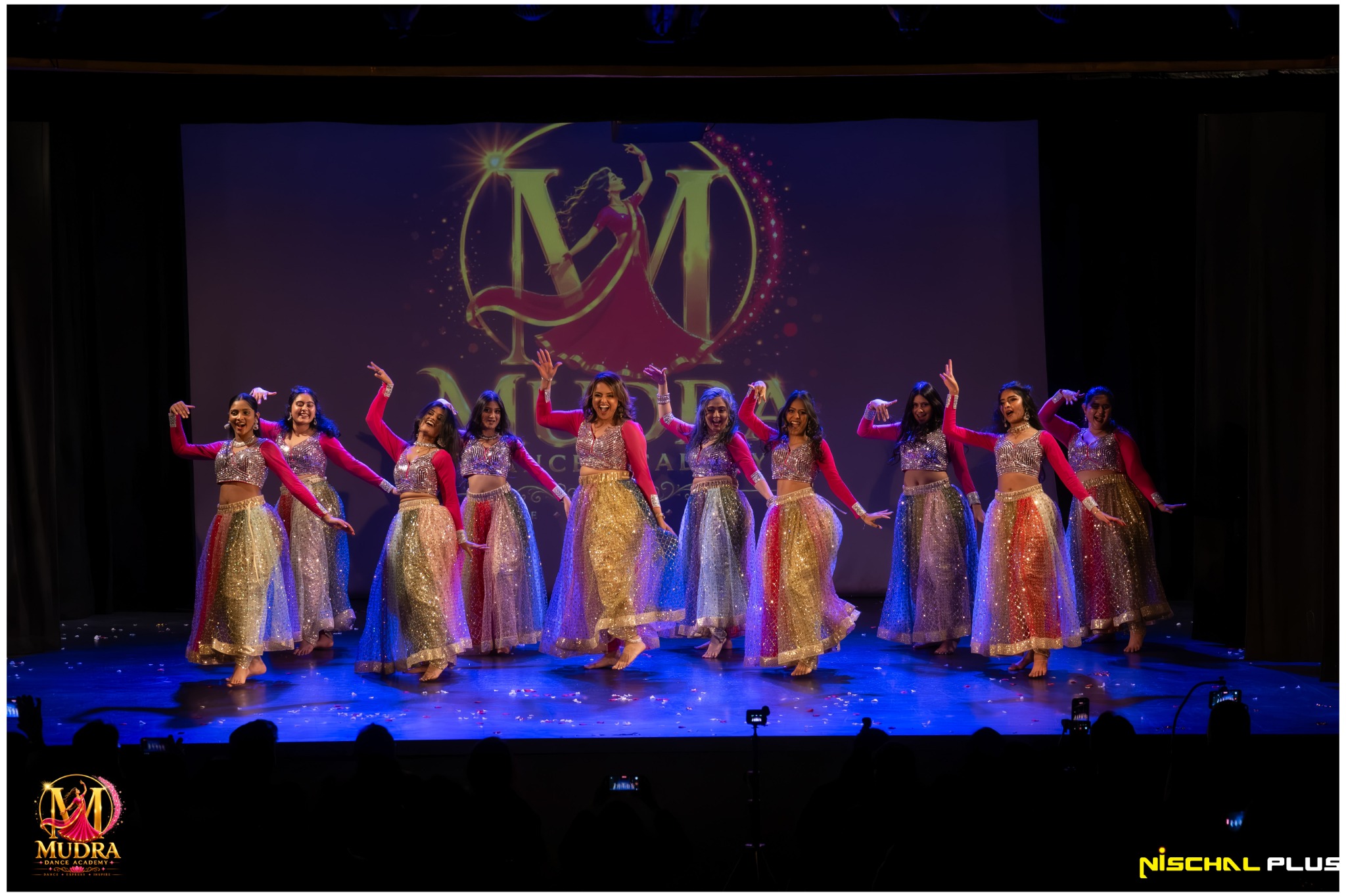 Large Mudra Dance Academy group performing in colourful costumes beneath the academy logo